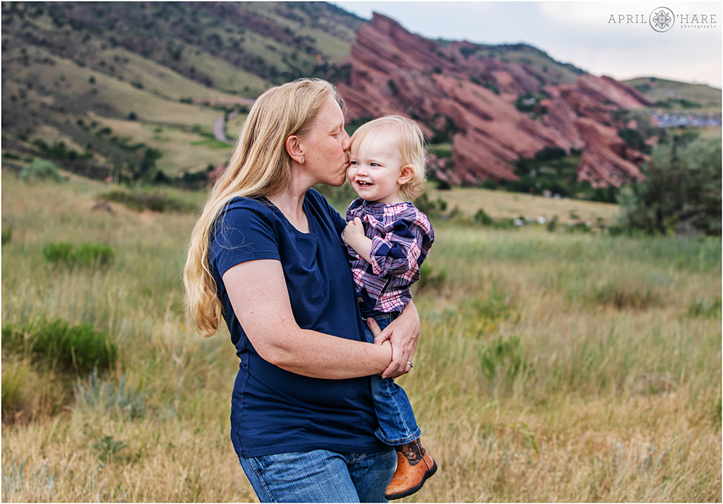 A mama kisses her sweet little girl during a family portrait session with Red Rocks Amphitheater in the backdrop at Mount Falcon in Colorado