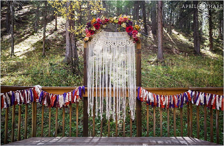 Colorado Boho Wedding Ceremony Decor with fall color jewel toned floral arch and ribbon garland and macrame backdrop in the woods