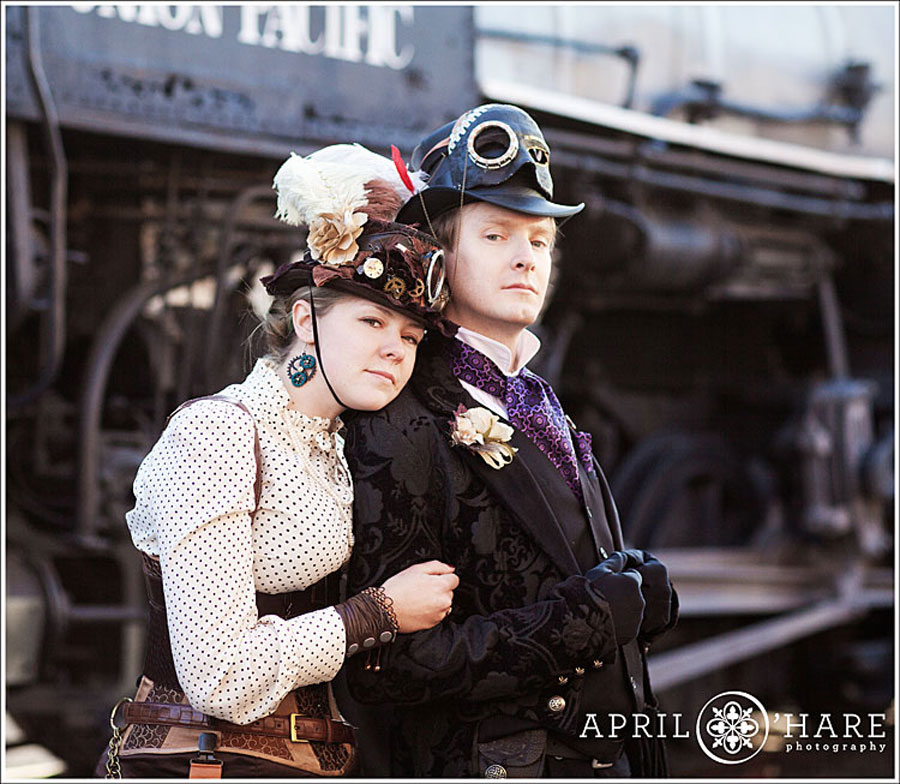 A cute couple dressed in old fashioned Steampunk Victorian clothing pose together in front of an old fashioned black railroad train car at the Colorado Railroad Museum in Golden Colorado during their winter engagement session with April O'Hare Photography based in Denver Colorado