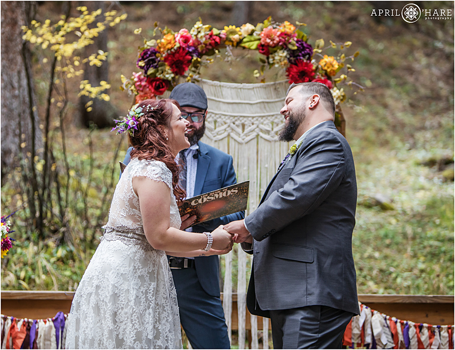Adorable moment at a Colorado Boho Wedding with pretty fall color floral arch and macrame wedding ceremony backdrop in the woods