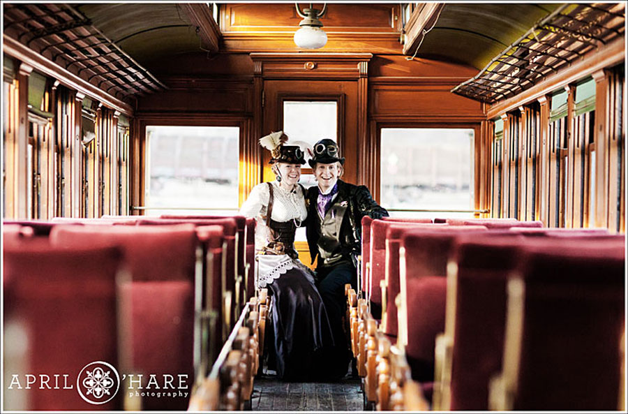 A cute couple dressed in old fashioned Steampunk Victorian clothing pose together on the inside of an old fashioned passenger railroad train car at the Colorado Railroad Museum in Golden Colorado during their winter engagement session with April O'Hare Photography based in Denver Colorado