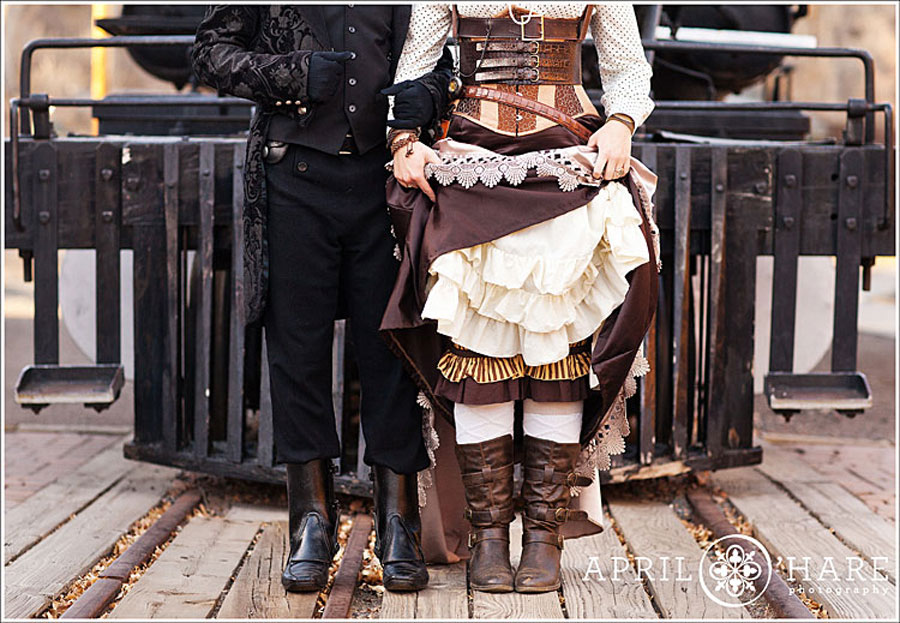 A cute couple dressed in old fashioned Steampunk Victorian clothing pose separately on in front of a cattle catcher of an old fashioned railroad train car at the Colorado Railroad Museum in Golden Colorado during their winter engagement photography session with April O'Hare Photography based in Denver Colorado.
