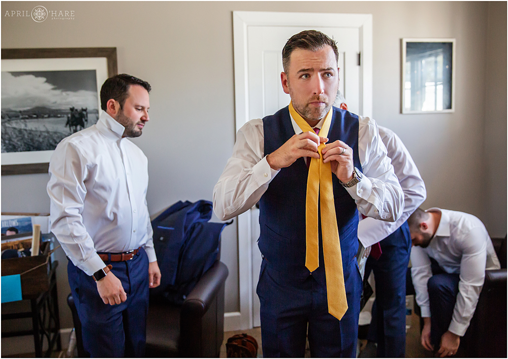 Groomsman helps groom with his yellow tie at The Barn at Raccoon Creek in Denver