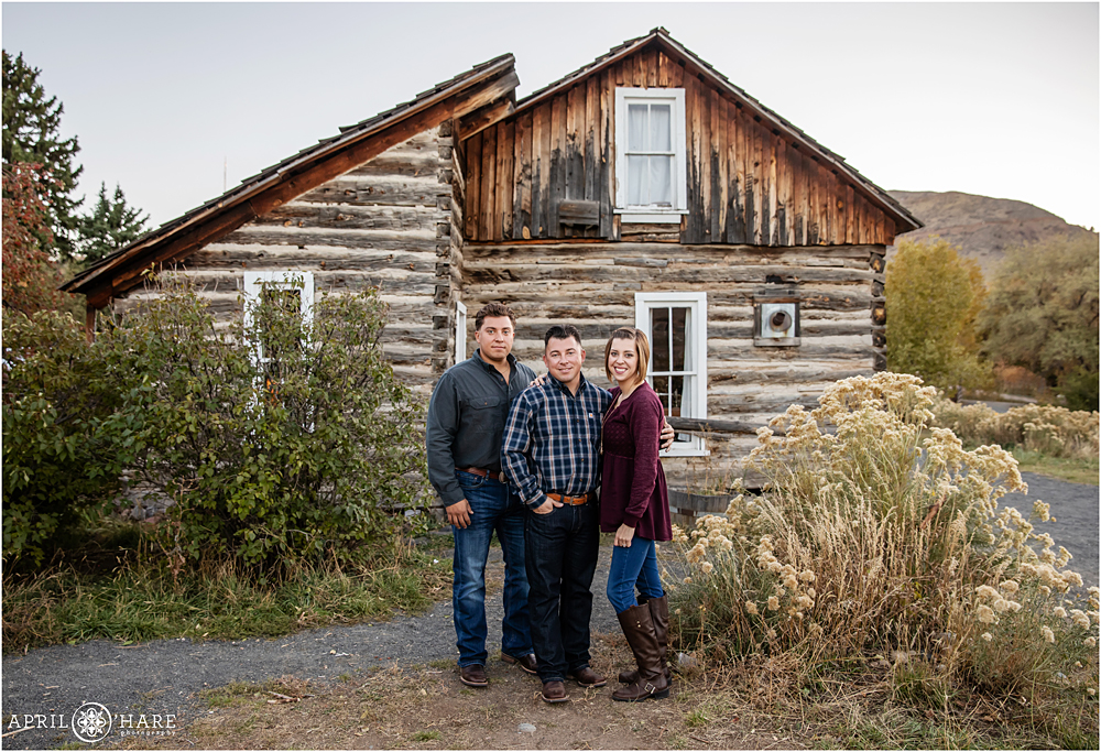 Adult siblings pose for a full length photo with a rustic cabin at the Clear Creek History Park in Golden Colorado
