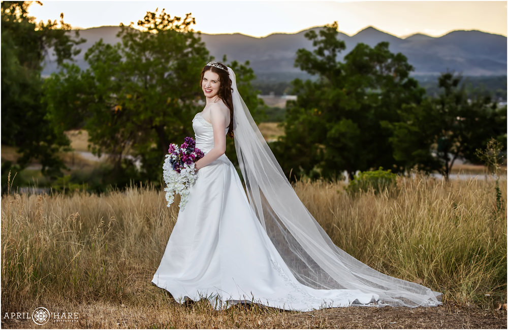 Bride wearing a tiara and holding purple and white bridal bouquet and long veil poses in front of a mountain view at Flyin' B Park