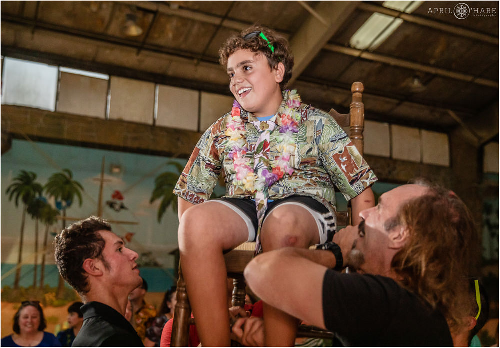 Bar mitzvah boy wearing hawaiian shirt at his jungle themed bar mitzvah party lifted in air for horah chair dance at The Island in Aurora CO