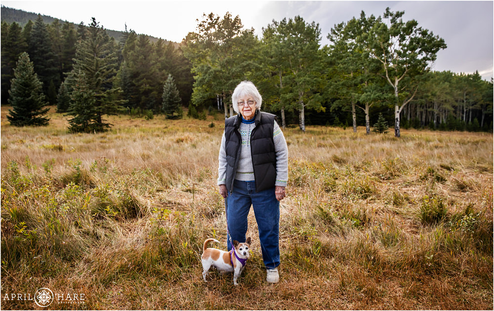 An older woman poses for a cute photo with her little dog while standing in a Colorado mountain meadow in Evergreen
