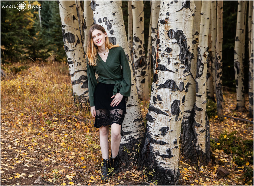 Senior girl wearing green leans against aspen trees for her Colorado yearbook photography session in Evergreen