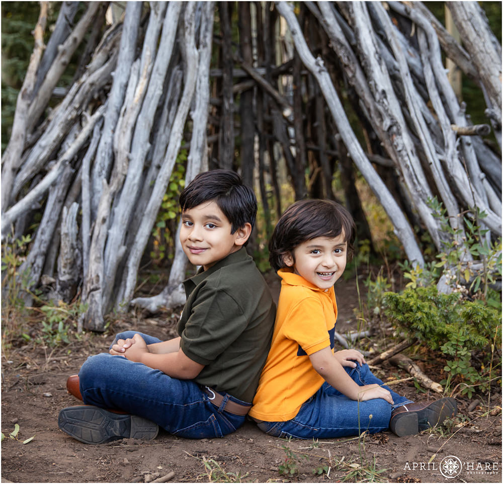 Two brothers sit on the ground back to back with wood tipi behind them in Colorado