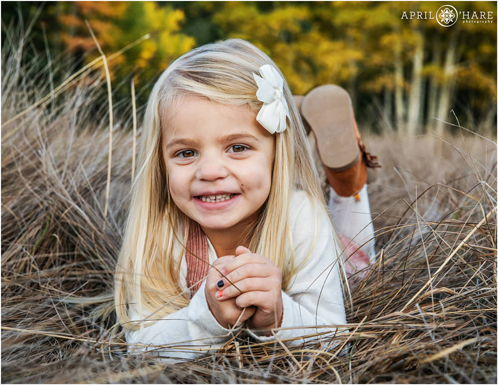 Adorable little blonde girl lying on the tall grass with pretty autumn tree backdrop on Squaw Pass Road family photo session