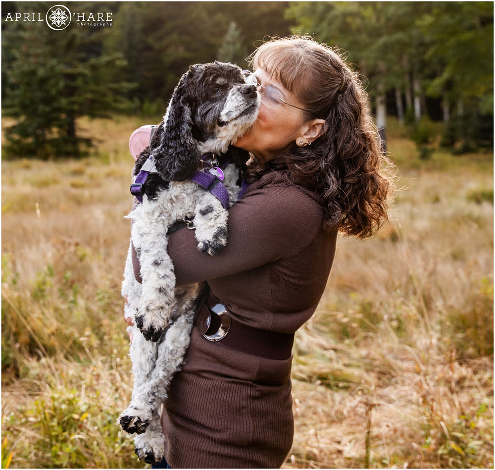 A woman kisses her older dog as she snuggles him during her family photography session in a mountain meadow in Colorado
