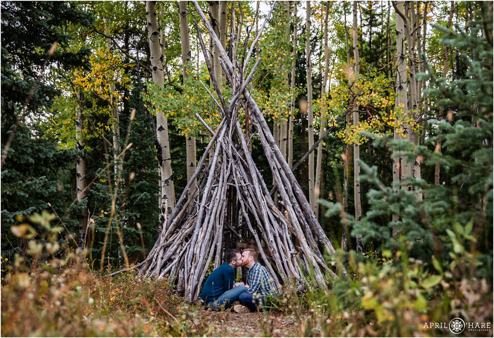 Romantic gay engagement photo two men seated on the ground lean toward each other with a pretty fall color backdrop in front of a wood tipi structure