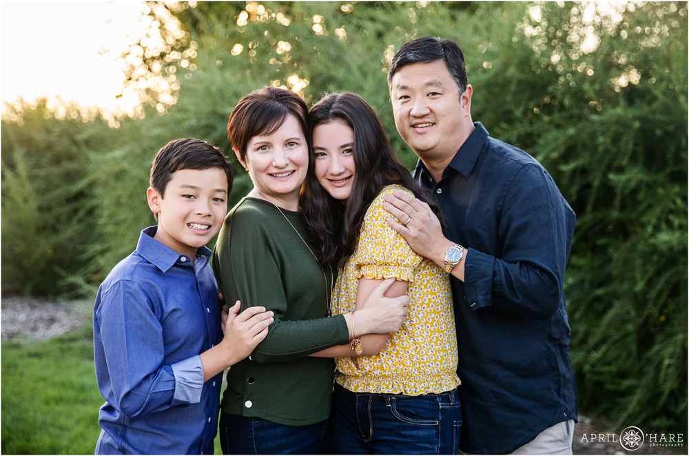 Family of four snuggle up together for their portraits in front of greenery at City Park in Denver Colorado