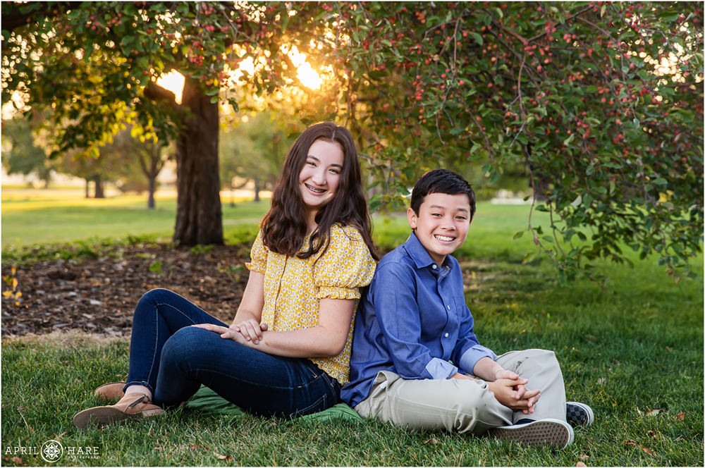Brother and sister sit back to back in front of red berry tree at a public park in Denver CO