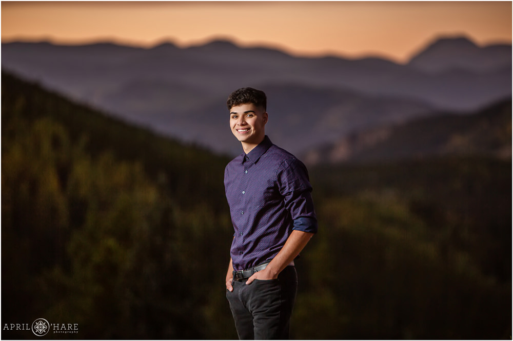 A young man poses for a senior photo in front of a beautiful sunset mountain backdrop on Squaw Pass Road in Evergreen