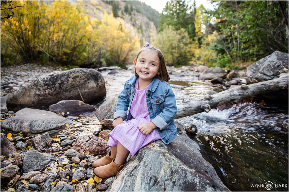 A cute girl wearing lilac dress with boots and jean jacket poses next to South Clear Creek in the mountains near Georgetown, CO