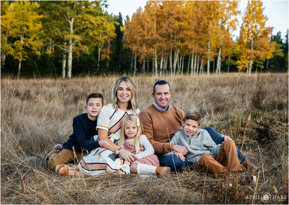 A family of five wearing burnt orange, navy, and peach sit in a pretty autumn colored mountain meadow in Colorado