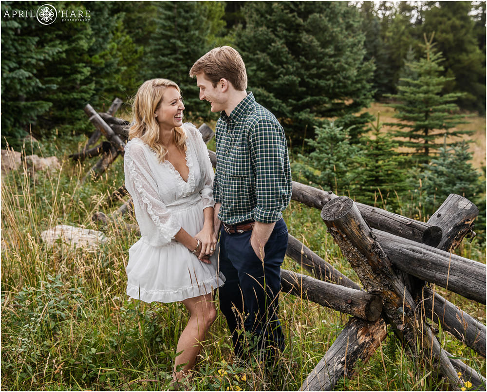 Young couple laugh together at their Colorado engagement session with rustic wood fence