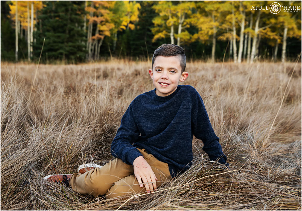 Boy wearing navy blue sweater and tan pants sits in a tall grassy autumn colored meadow in Colorado