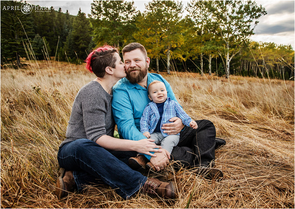 Mom and dad kiss while their baby smiles in a pretty autumn mountain meadow