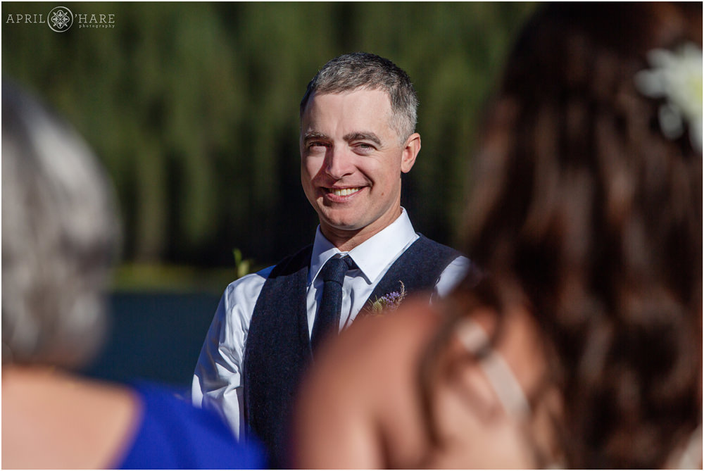 Groom smiles at his bride as she walks down the aisle toward him at their sunny outdoor Vail wedding