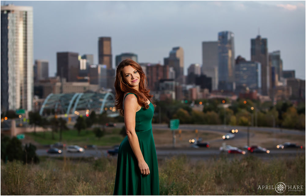 A beautiful red head woman wearing a long flowing sleeveless green dress photographed at dusk in front of Denver city skyline