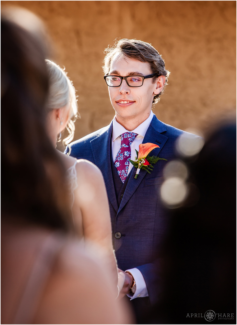 Groom wearing glasses in a blue suit with orange boutonniere at outdoor wedding ceremony in Courtyard at Villa Parker in Colorado
