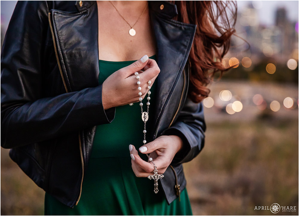 A close up detail of a woman with red hair holding a rosary in front of Denver city skyline at sunset