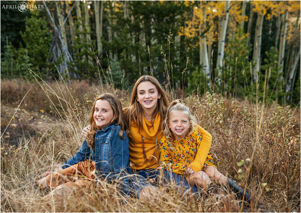 Three cute sisters sit in a mountain field with a woods backdrop on Squaw Pass Road during Autumn in Colorado