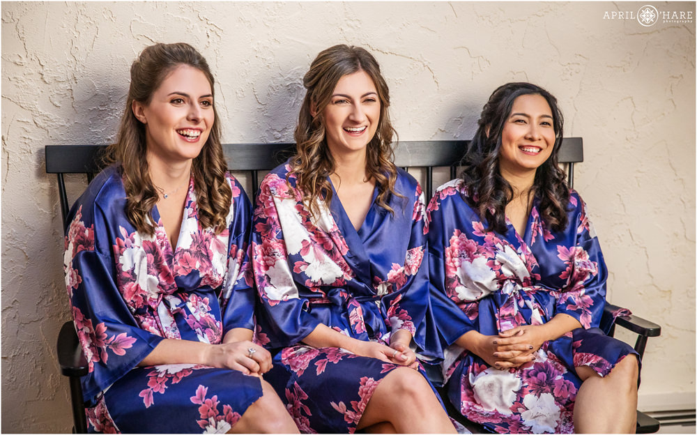 Bridesmaids all wearing matching purple silk floral robes sit on a bench together at Villa Parker in Colorado