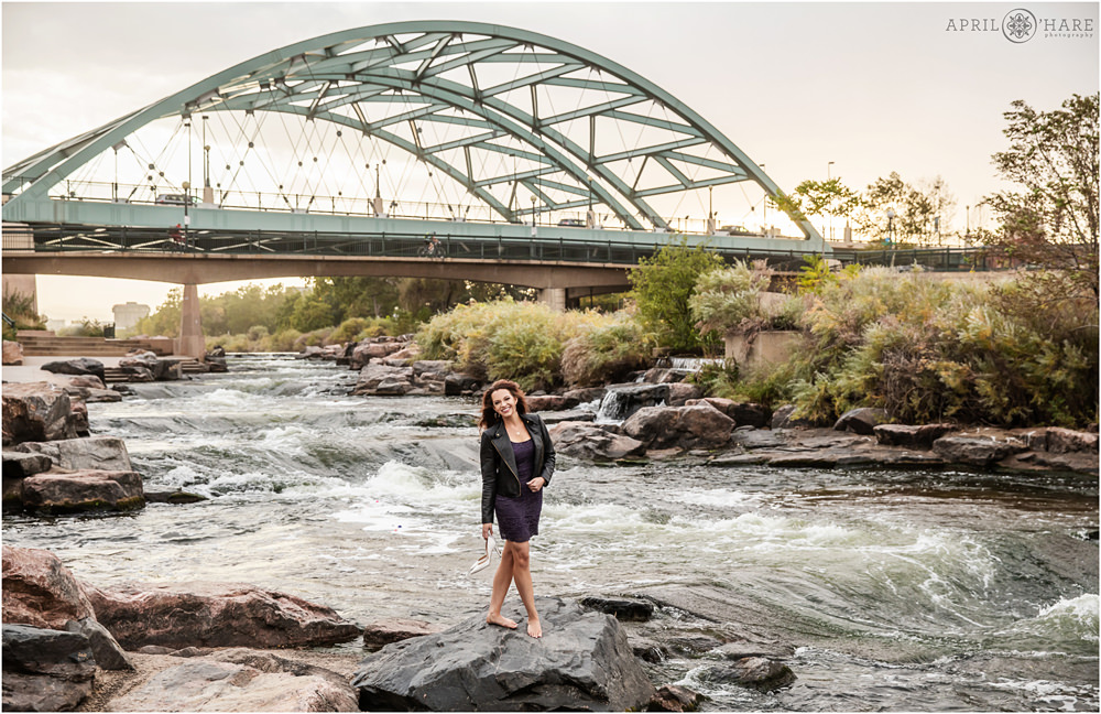 A woman holding her white heels in her hand poses on the rocks next to South Platte River with the Speer Bridge in the backdrop in Denver