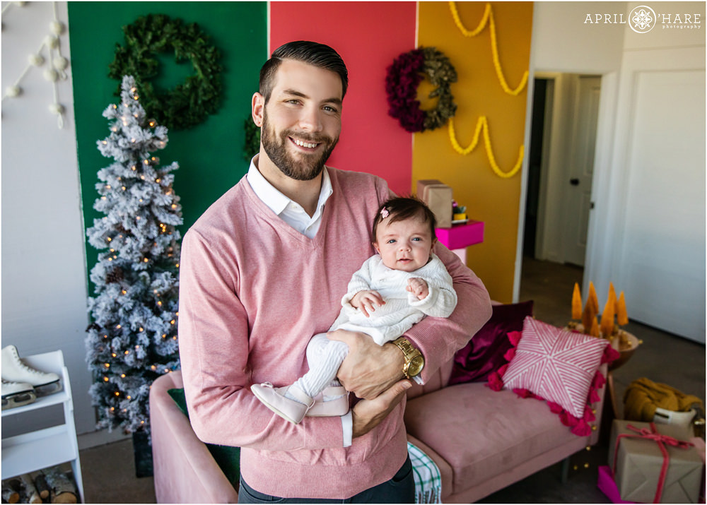 A dad wearing pink poses with his sweet new baby at a bright colorful holiday family photography session in Denver