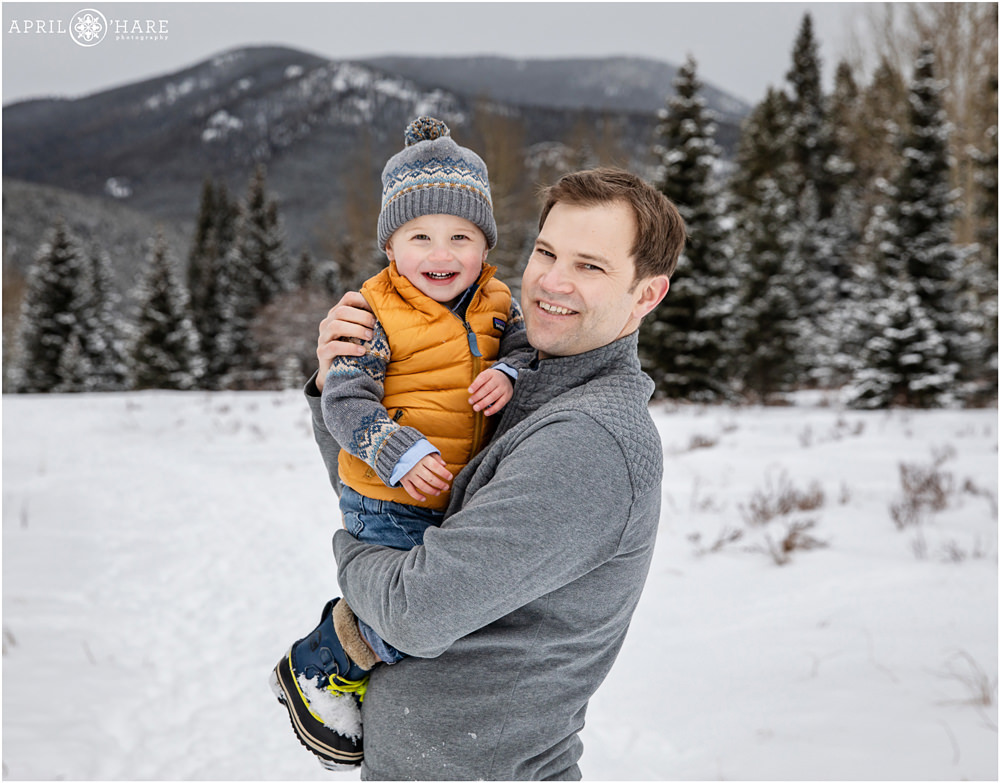 Dad and son wearing gray and yellow pose for family photos during winter in a mountain forest in Colorado