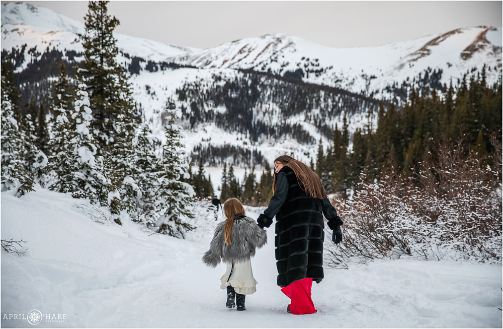 Mom with long brunette hair wearing a black coat and full length red dress walks hand in hand with her daughter through a snowy winter landscape at Mayflower Gulch Trailhead