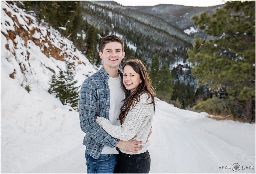 Cute couple pose for a portrait on a snowy winter road with evergreens dotting the mountains behind them