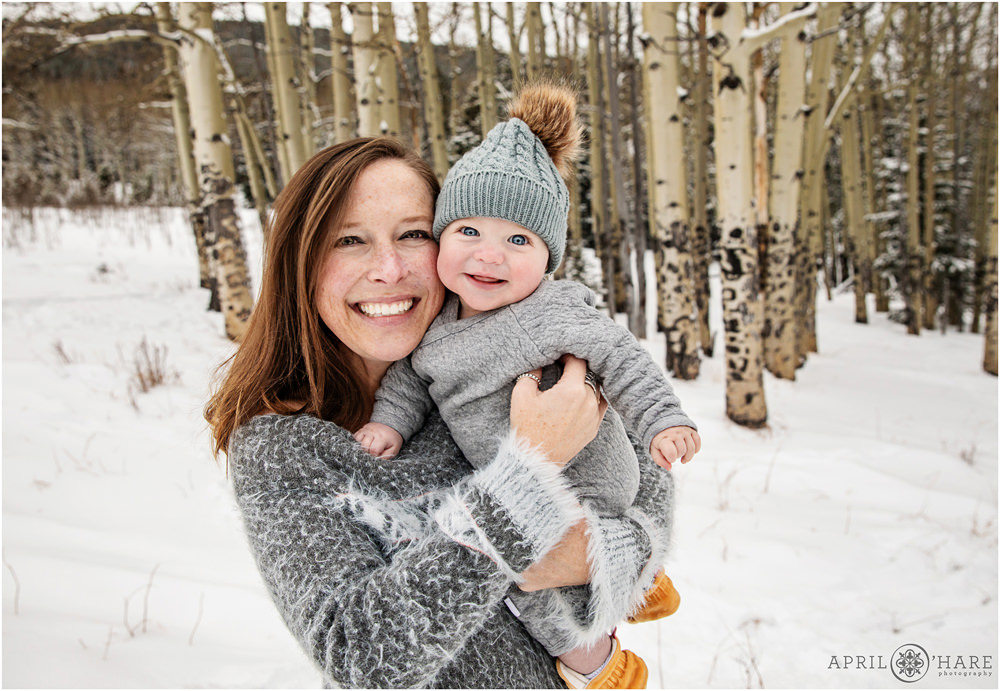 A mom holding her smiling 6 month old baby both wearing gray with aspen tree backdrop in a mountain meadow during winter