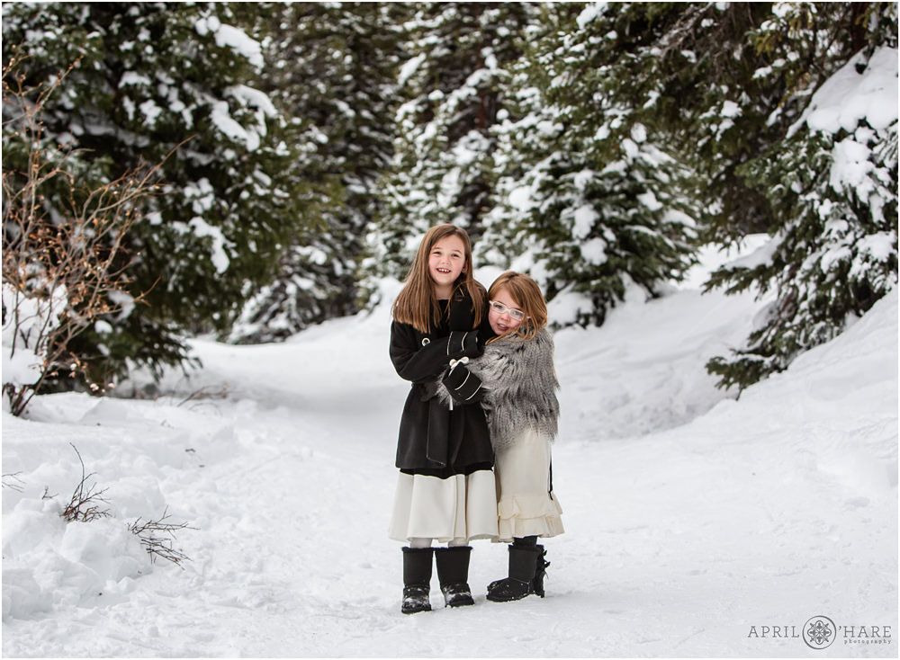 Two sisters wearing cute dresses and fancy coats hug on a woodsy snow covered path at Mayflower Gulch near Leadville