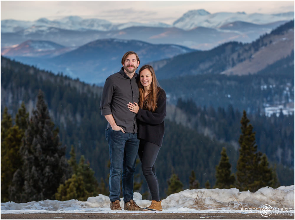 Young couple wearing gray & black pose in front of snowy mountain backdrop in Colorado