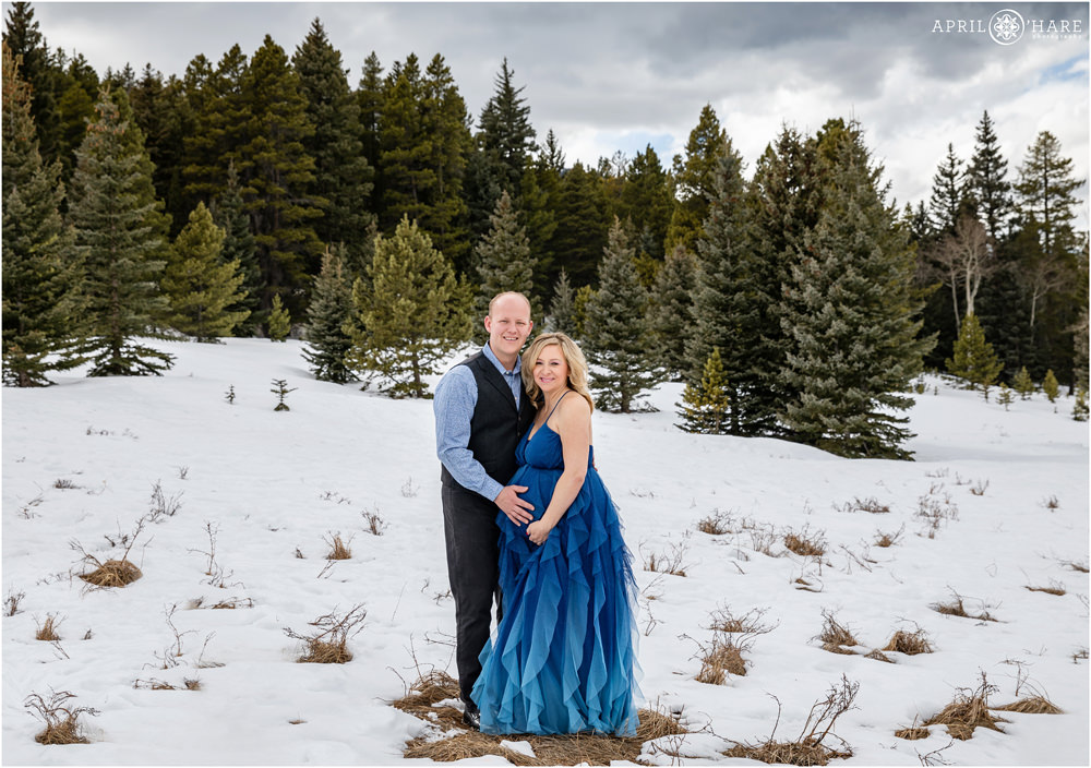 Cute couple pose for a maternity photo in a pretty snowy landscape with evergreen tree backdrop in Evergreen Colorado
