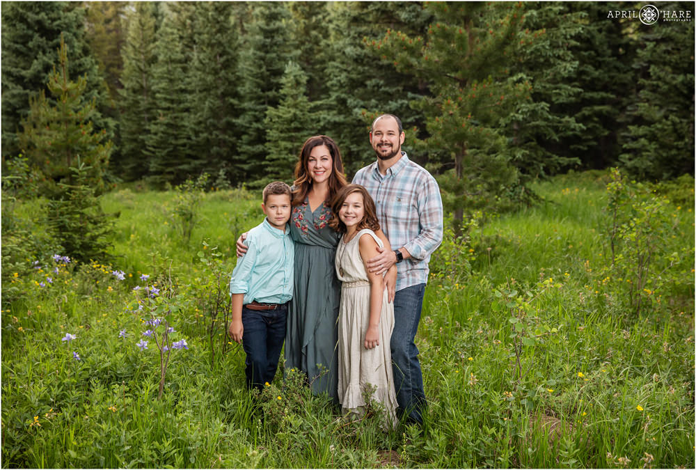 A cute family of four wearing shades of blue and teal and cream pose in a mountain forest with wildflowers in Colorado