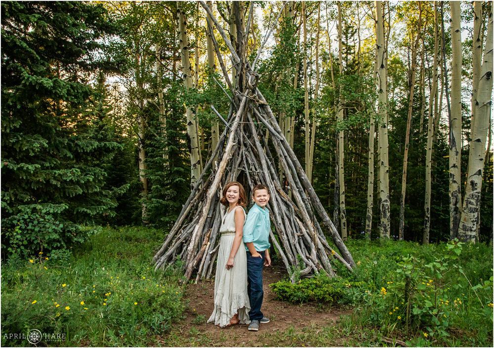 Brother and sister laugh together in front of a cool wood tipi structure in a Colorado Aspen tree forest in Evergreen