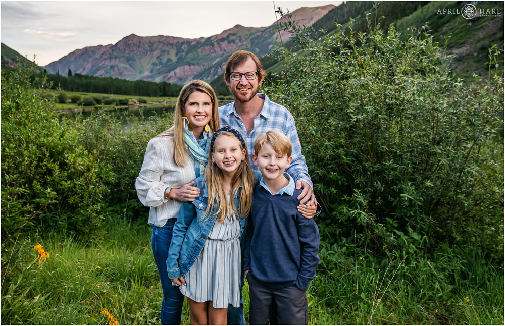 Beautiful family of 4 wearing blue pose in front of the green and red mountain backdrop at Maroon Lake