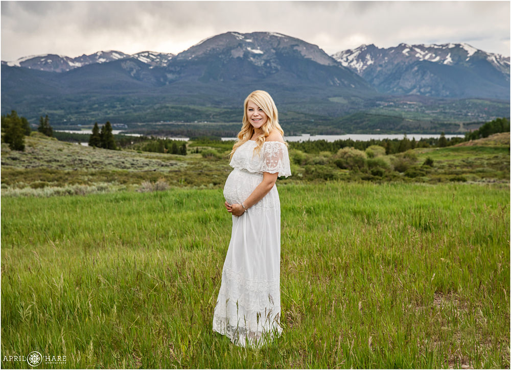 A beautiful blonde pregnant woman poses for a full length maternity portrait in her white boho gown in front of a mountain view in Summit County Colorado