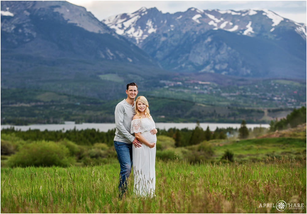 A good looking couple pose for their maternity photos in front of a dramatic mountain view in a grassy field during summer in Summit County Colorado