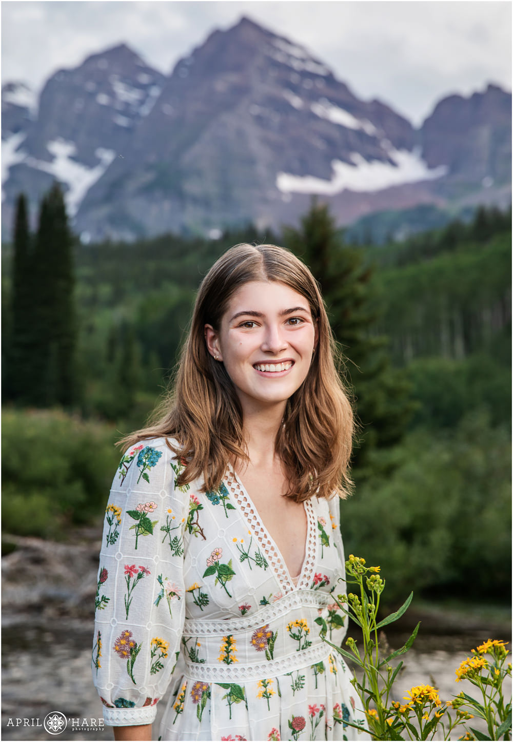 A young woman wearing a pretty white floral print dress poses for an individual portrait with a mountain backdrop near Snowmass Village Colorado