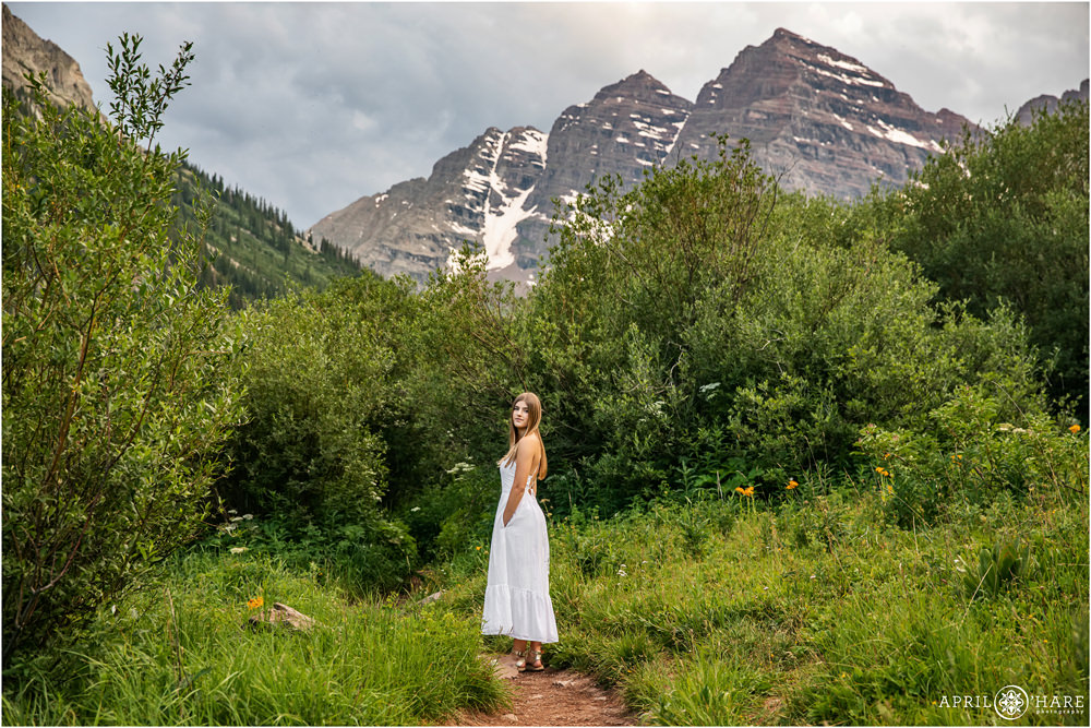 High school girl with long straight hair wearing a full length white dress poses on path at Maroon Bells