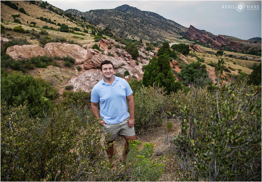 A high school senior boy wearing a striped light blue shirt and khaki shorts poses with beautiful scenery at East Mount Falcon Trailhead