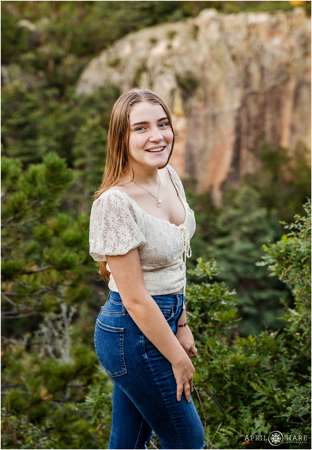 Teen girl with long blonde hair wearing a cream top and jeans poses in front of a pretty backdrop at Lovers Leap in Colorado