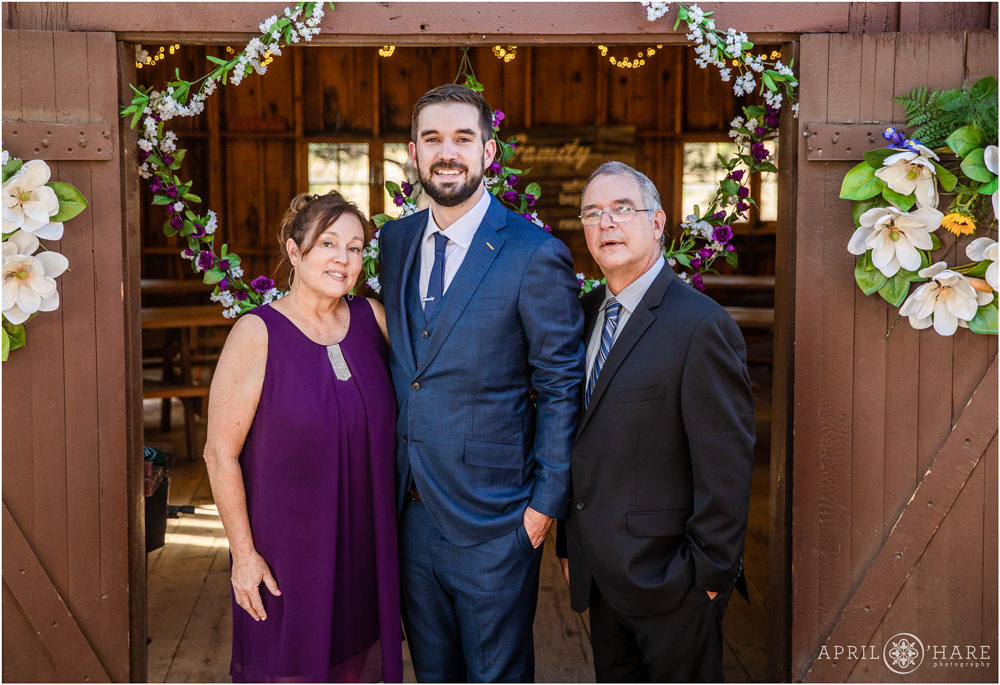 Groom poses with his mom and dad in front of a rustic barn at his Colorado wedding