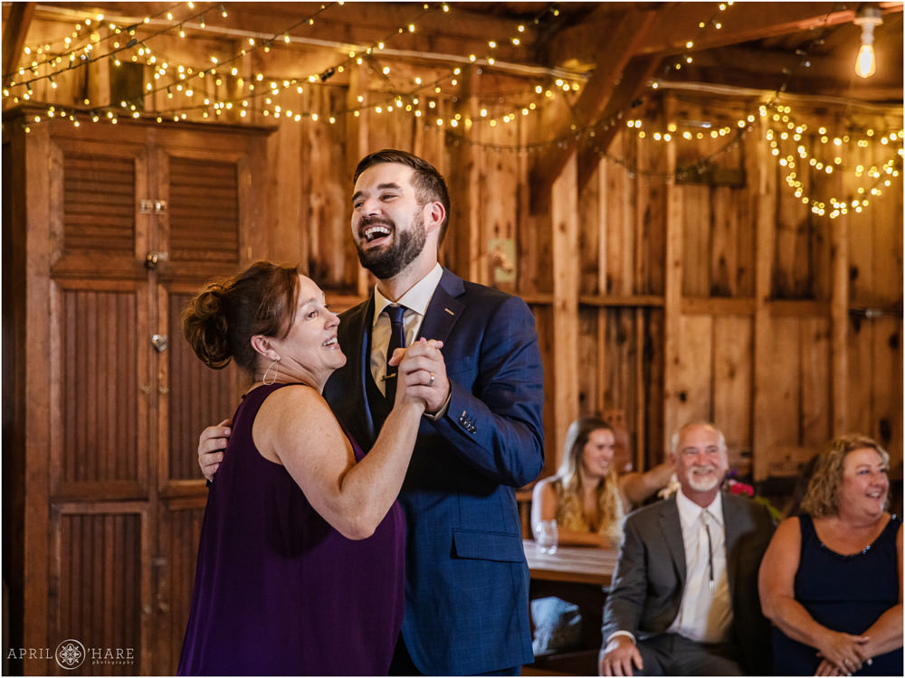 Groom laughs with his mom as they dance on his wedding day inside a small rustic barn in Lookout Mountain in Colorado